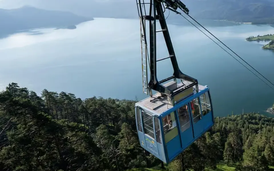 Seilbahnkabine über bewaldetem Hang mit See und Bergen im Hintergrund